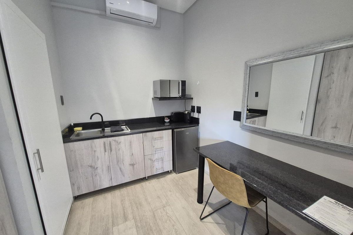 A compact kitchenette featuring light wood-grain cabinetry, dark stone countertops, a stainless steel sink, and a wall-mounted air conditioning unit.