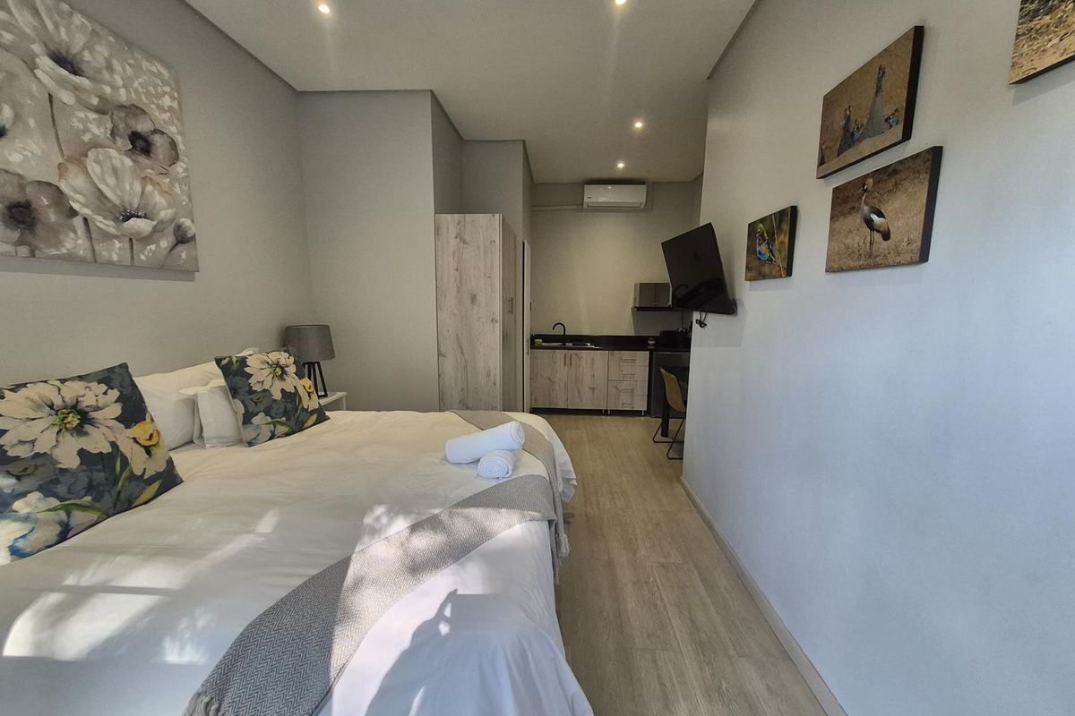 A studio-style bedroom featuring light wood-look flooring, recessed ceiling lighting, a wall-mounted air conditioning unit, and a compact kitchenette area with built-in cabinetry.