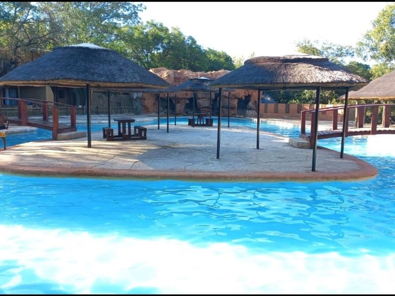 Outdoor swimming pool area featuring a central paved island with thatched umbrellas, connected by wooden footbridges and accented by a natural rock feature.