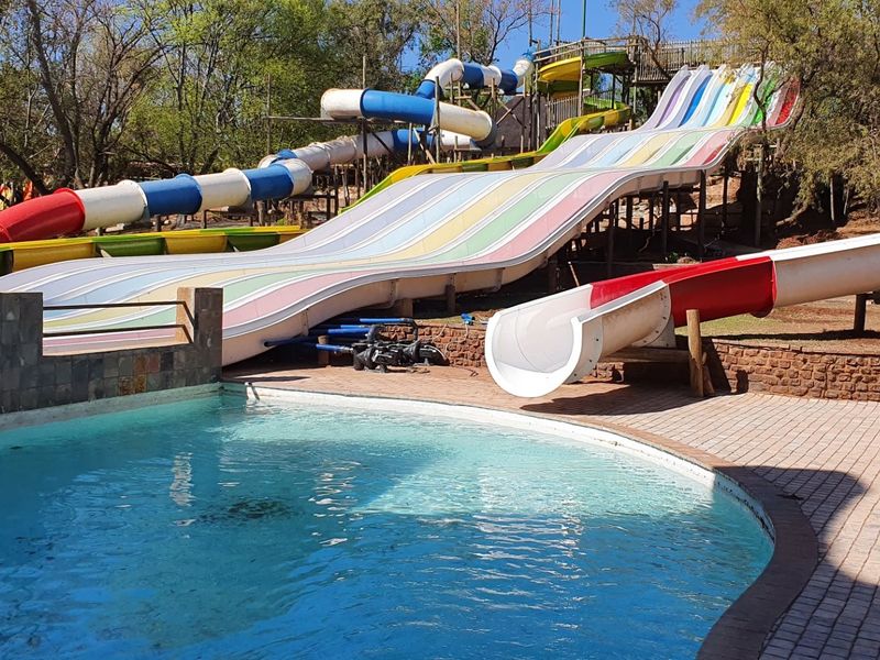Outdoor pool area featuring multiple large water slides, a paved deck, and stone retaining walls in a park-like setting.