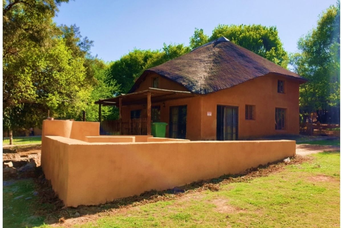 A rustic building featuring a traditional thatch roof, earthen-toned exterior walls, and a covered patio area set within a landscaped garden.