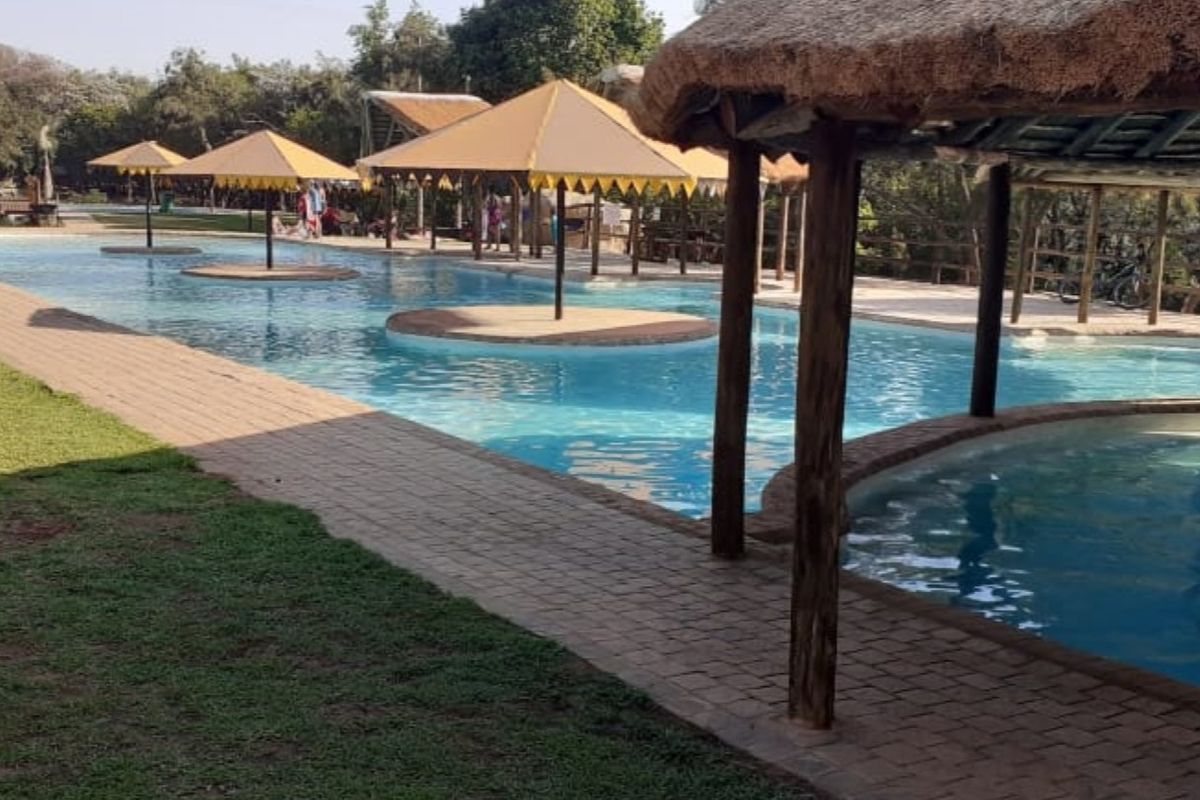 Outdoor swimming pool area featuring multiple thatched shade structures, paved walkways, and landscaped surroundings, ideal for recreational use.