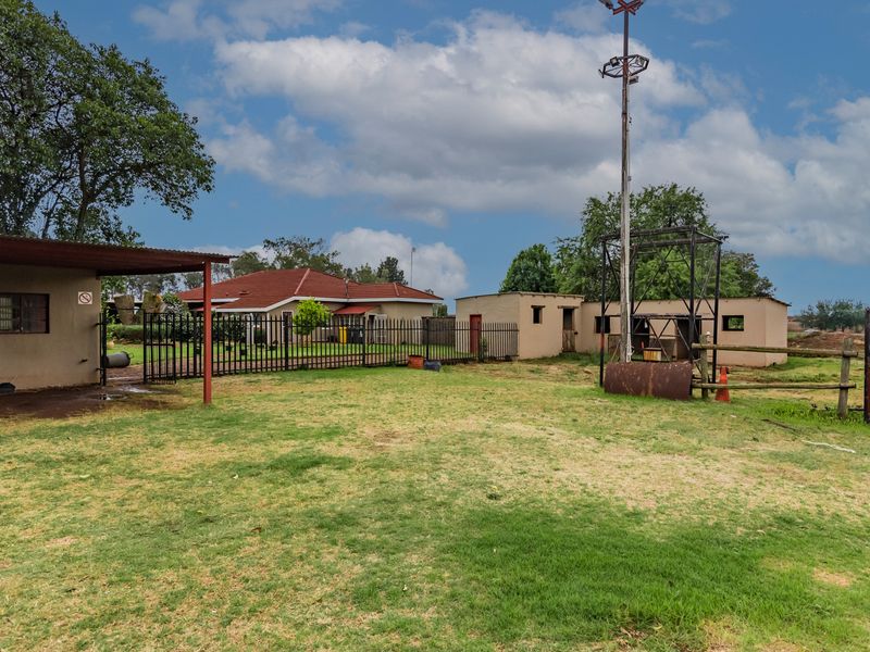 Rear exterior view of the small holding featuring a large open lawn, metal fencing, a carport, and various utility structures.