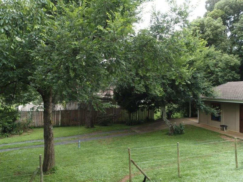 A spacious backyard garden featuring a lush lawn, mature trees, a paved walkway, and a section of the house with a tiled roof.