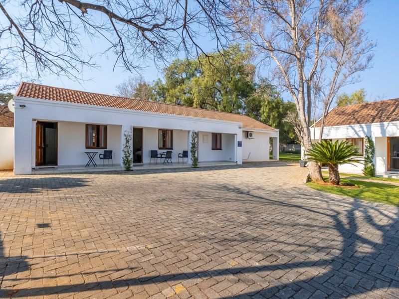 A well-maintained guest house exterior featuring a spacious paved driveway, covered patio areas, and established garden surroundings under a clear sky.