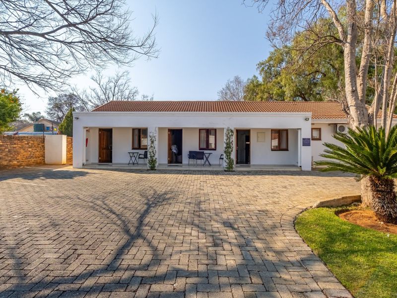 A well-maintained guest house exterior featuring a paved driveway, covered patio area, and established garden surroundings.