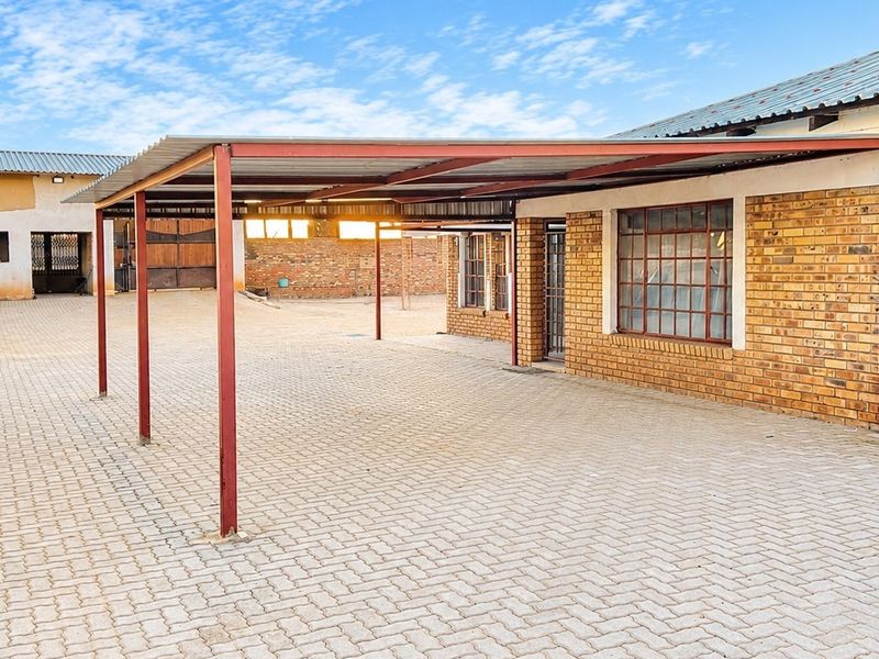 A spacious paved driveway featuring a large metal carport structure attached to a brick-faced building with steel-framed windows.