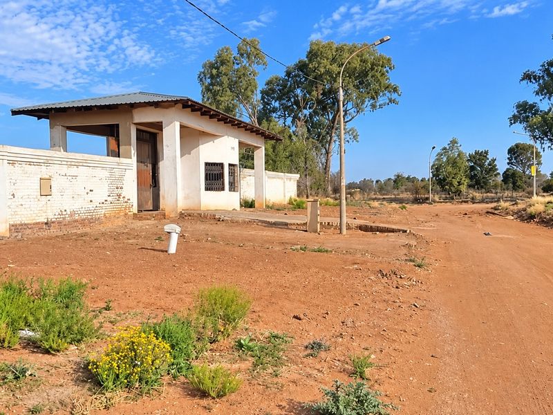 Exterior view of a property entrance featuring a security gatehouse, perimeter wall, and unpaved access road in a rural or semi-developed setting.