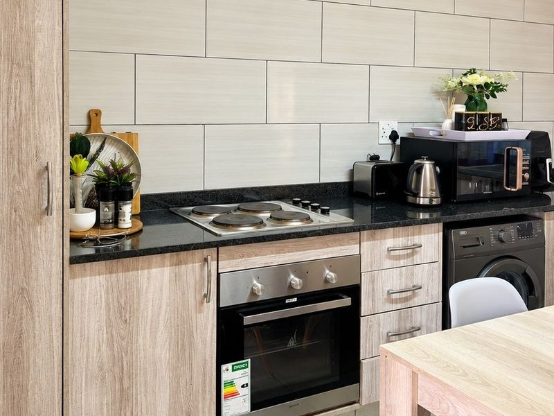 Modern kitchen featuring light wood-grain cabinetry, granite countertops, a tiled backsplash, and integrated appliances including an oven, hob, and washing machine.
