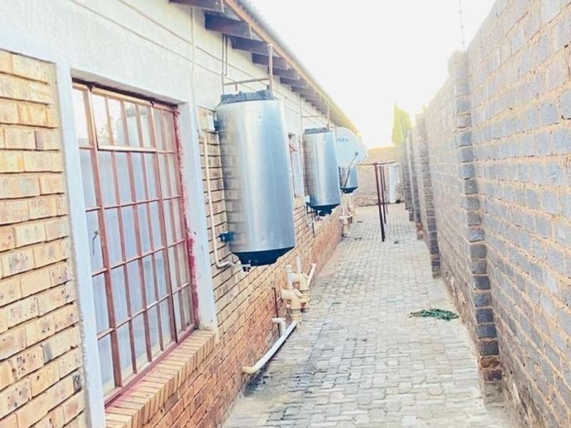 A narrow rear exterior walkway featuring exposed brick walls, paved flooring, and multiple wall-mounted electric water geysers.
