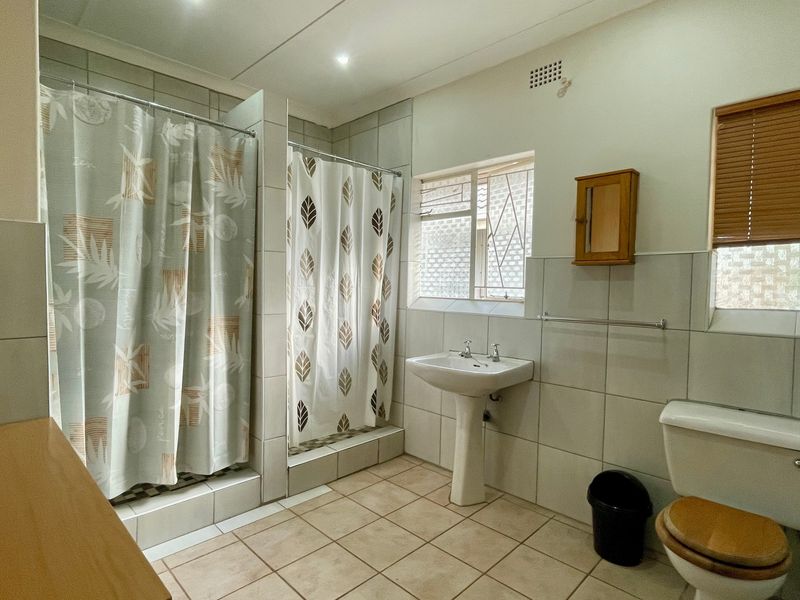 A functional bathroom featuring a pedestal washbasin, toilet, and a tiled shower area with neutral wall and floor tiles.
