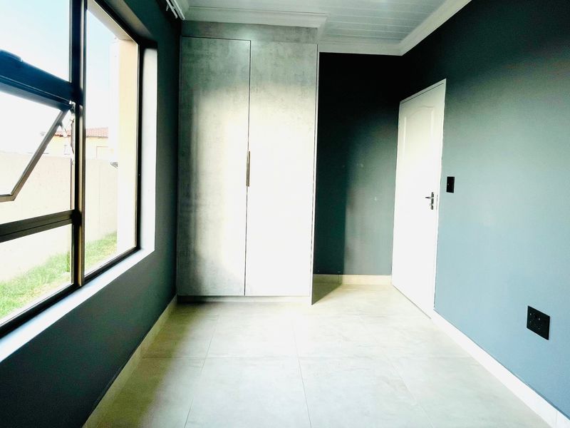 A bright, empty bedroom featuring modern tiled flooring, a built-in wardrobe, and dark accent walls with large windows providing natural light.