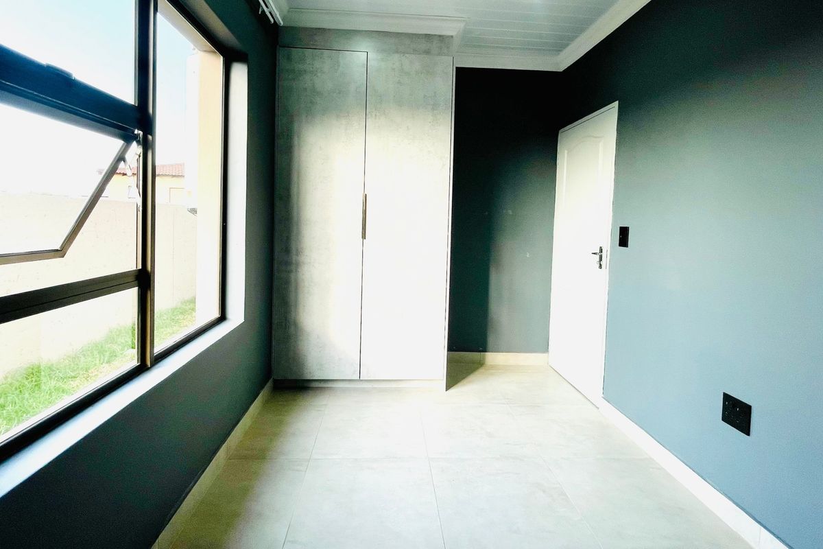 A bright, empty bedroom featuring modern tiled flooring, a built-in wardrobe, and dark accent walls with large windows providing natural light.