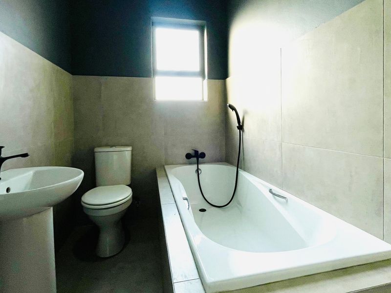 A modern bathroom featuring a pedestal basin, a standard toilet, and a built-in bathtub with a handheld shower mixer, finished with neutral wall tiles.