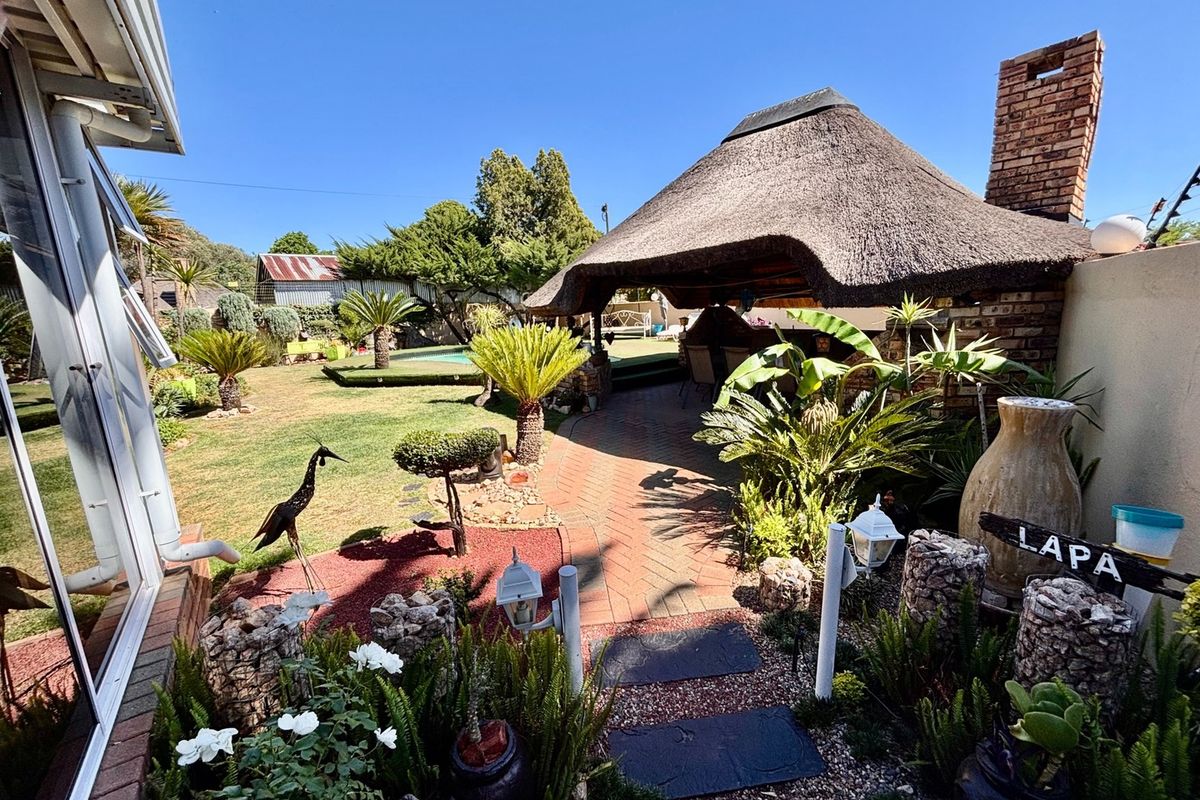 A well-maintained rear garden featuring a traditional thatched lapa with a brick chimney, paved pathways, and a swimming pool in the background.