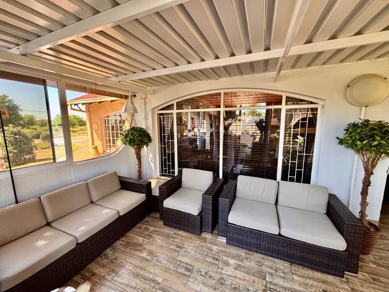 A covered patio area featuring tiled flooring, a corrugated metal roof, and large windows with security bars, providing a comfortable outdoor living space.