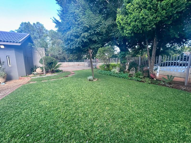 A well-maintained residential garden featuring a lush green lawn, mature trees, and secure perimeter fencing with a paved driveway area.