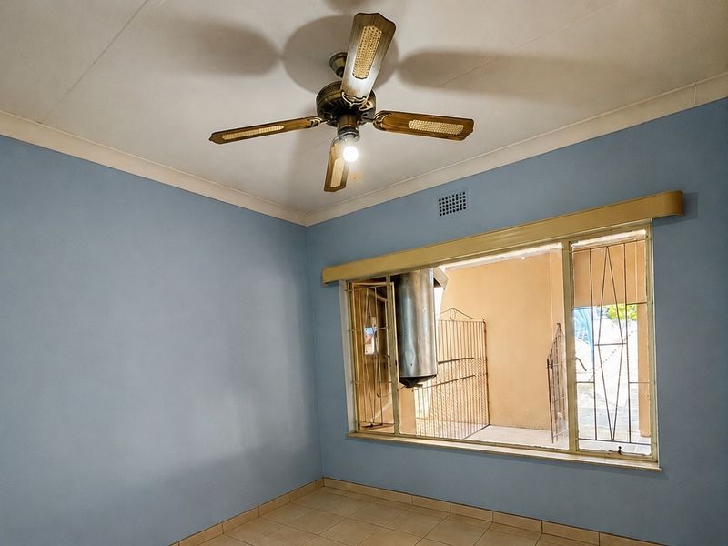 An empty bedroom featuring tiled flooring, built-in wooden cupboards, a ceiling fan, and large windows fitted with security bars.