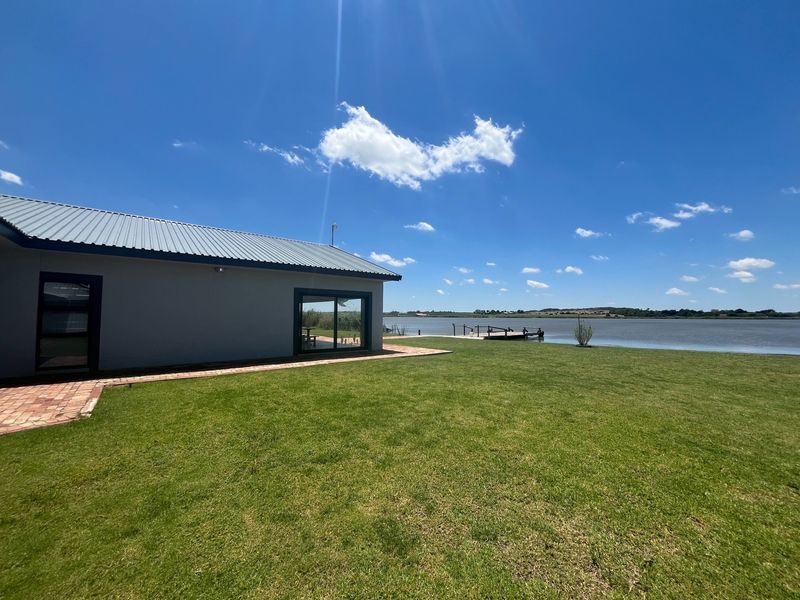 Rear view of the property featuring a metal roof, sliding glass doors, a brick walkway, and a spacious lawn overlooking the water and dock.