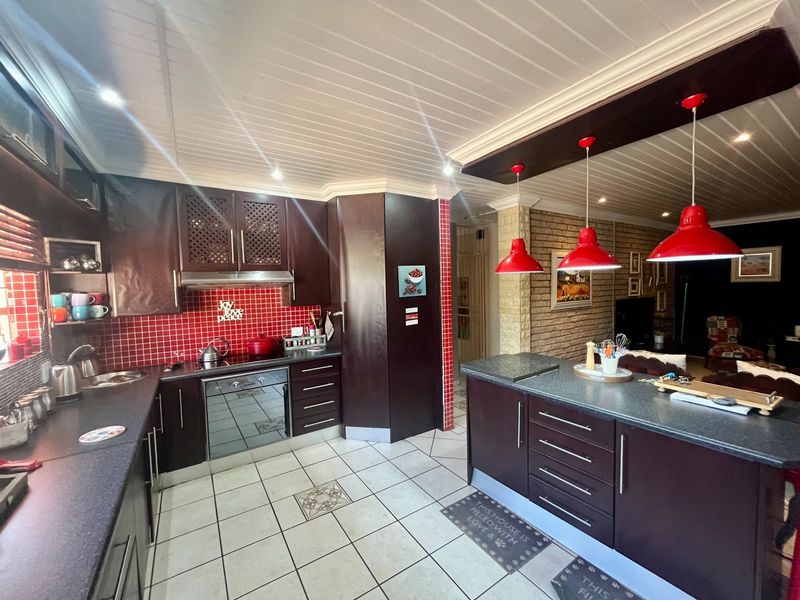 Open-plan kitchen featuring dark cabinetry, a central island with pendant lighting, red tiled backsplash, and tiled flooring, seamlessly connecting to the living area.