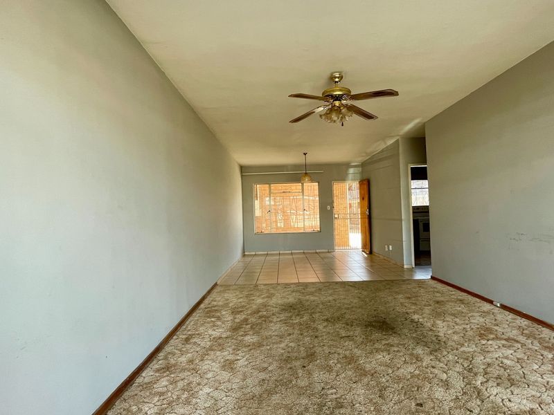 An empty open-plan living area featuring a combination of carpeted and tiled flooring, a ceiling fan, and access to a security gate.
