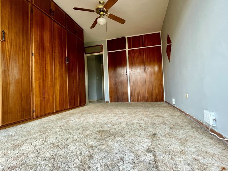 An empty bedroom featuring extensive built-in wooden cabinetry, a ceiling fan, and worn carpet flooring, offering a blank canvas for renovation.