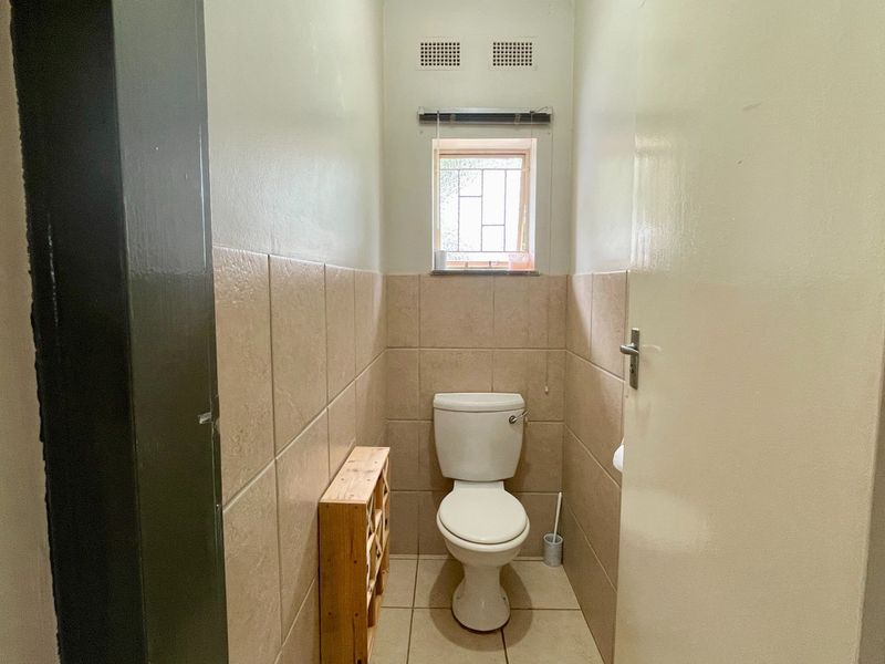A compact, functional guest toilet featuring neutral ceramic wall and floor tiling, a window with security bars, and ventilation grilles.