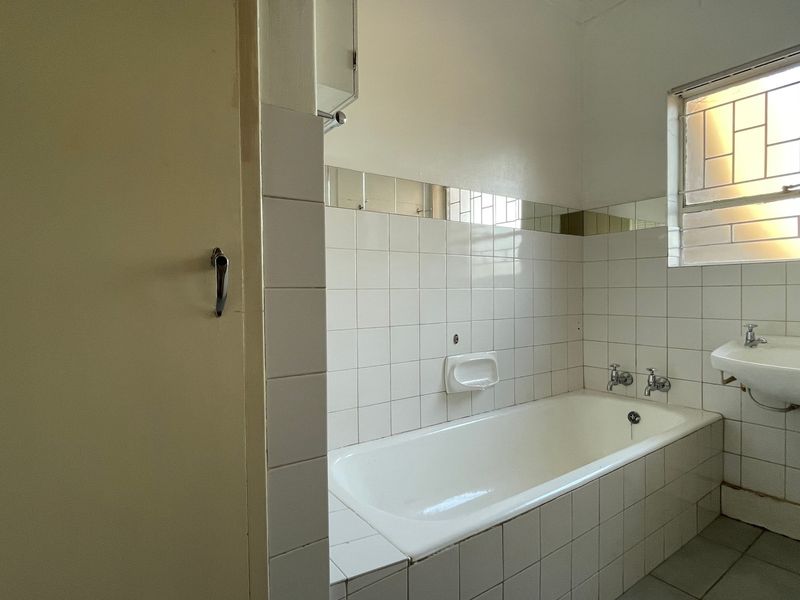 A functional bathroom featuring a built-in bathtub, wall-mounted basin, and white ceramic tiling, offering a blank canvas for modern renovation.