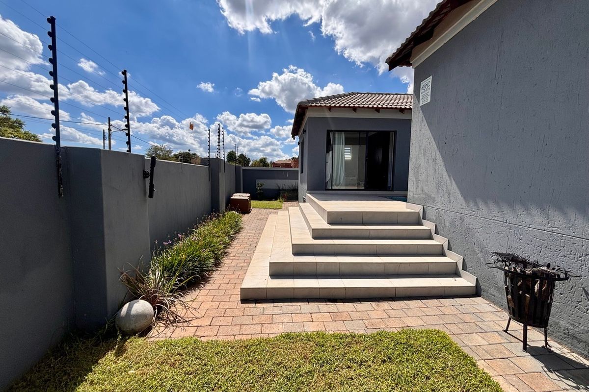 A well-maintained rear exterior featuring a paved walkway, tiled patio steps leading to a sliding glass door, and secure boundary walls with electric fencing.