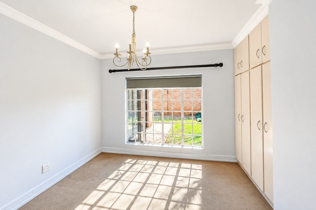 A bright, empty bedroom featuring neutral carpeted flooring, built-in cupboards, and a classic chandelier, with a view overlooking the rear garden.