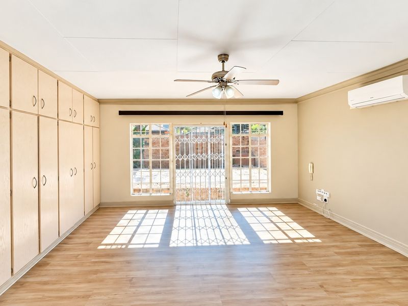 Spacious living area featuring light wood-finish built-in cupboards, laminate flooring, a ceiling fan, and a wall-mounted air conditioning unit with access to the rear patio.
