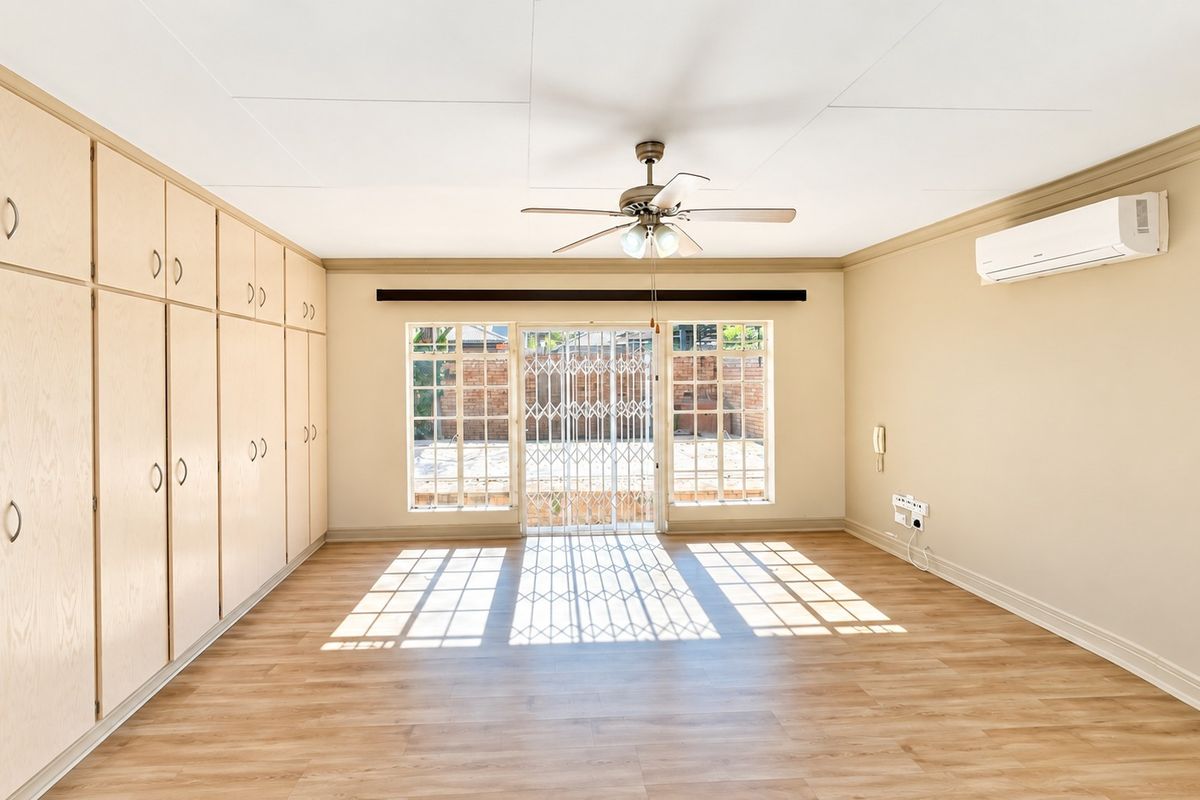 Spacious living area featuring light wood-finish built-in cupboards, laminate flooring, a ceiling fan, and a wall-mounted air conditioning unit with access to the rear patio.