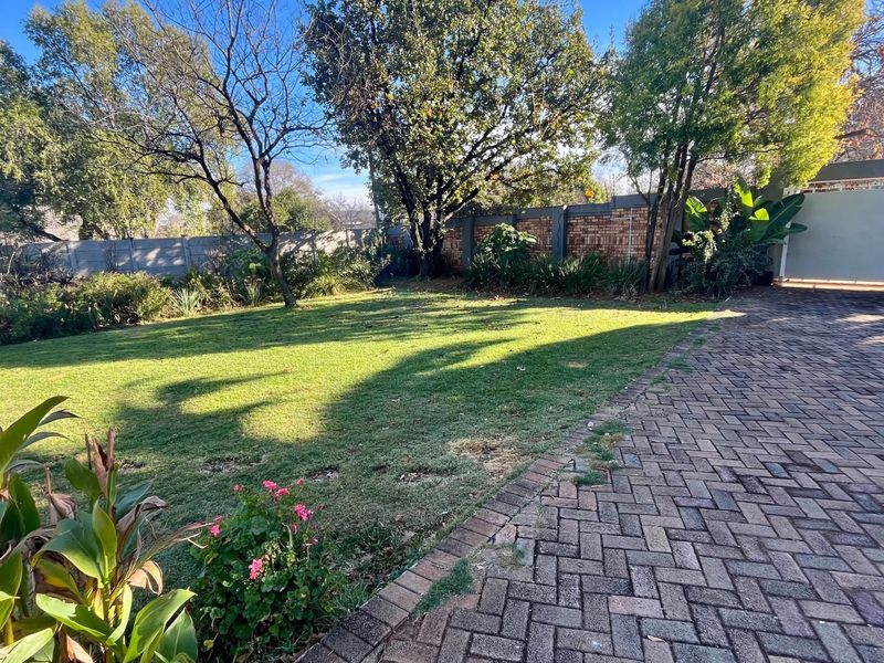 A spacious backyard featuring a well-maintained lawn, mature trees, and a paved driveway area enclosed by a brick boundary wall.