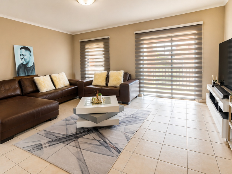 A well-maintained living room featuring neutral tiled flooring, modern roller blinds, and a sliding glass door providing natural light.