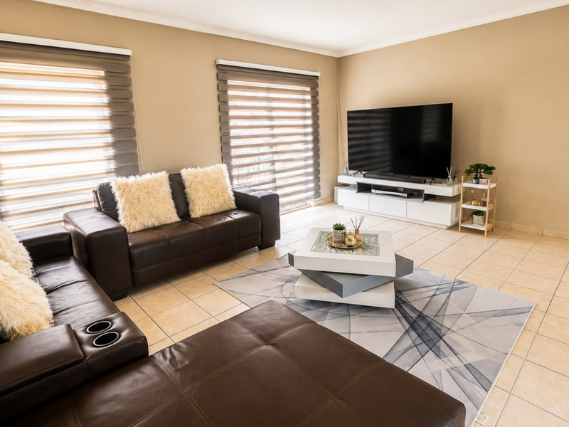 A well-maintained living room featuring neutral wall tones, tiled flooring, and dual roller blinds providing natural light control.
