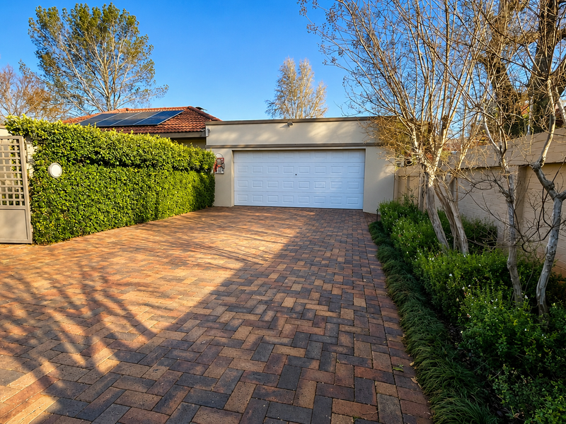 A well-maintained front exterior featuring a paved driveway, a secure garage door, and neatly landscaped boundary walls with established hedging.
