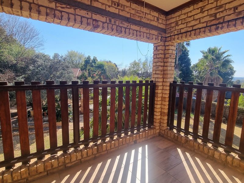 A covered balcony featuring exposed brick pillars, a timber balustrade, and tiled flooring, offering an open view of the surrounding garden area.