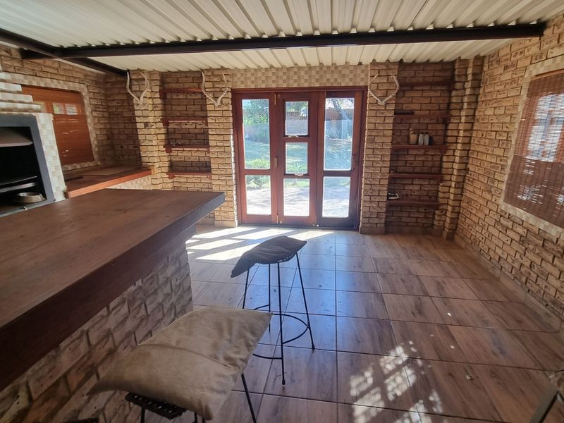 Covered patio area featuring exposed brick walls, a built-in braai, tiled flooring, and timber-framed glass doors leading to the garden.