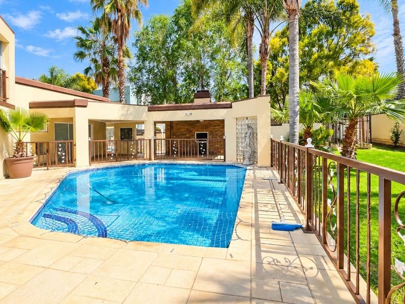 Outdoor pool area featuring a tiled deck, metal safety fencing, and a covered patio with a built-in braai and stone feature wall.