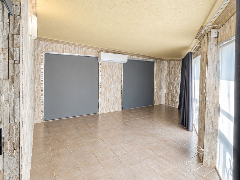 An empty living area featuring stone veneer wall cladding, ceramic floor tiles, and a split-system air conditioner, with large sliding glass doors and roller blinds.