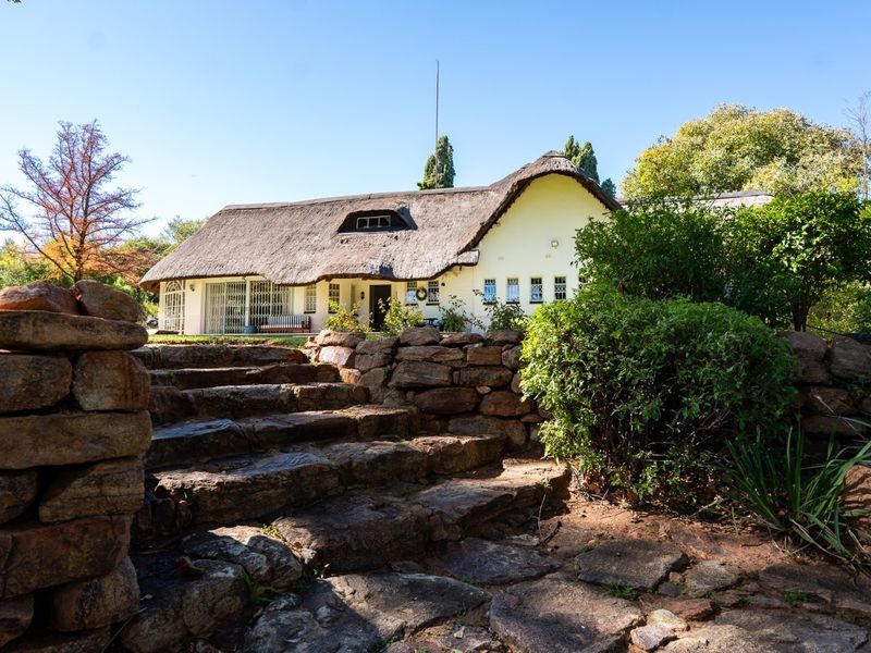 A charming thatched-roof house viewed from the rear garden, featuring natural stone retaining walls and a stone staircase leading to the residence.