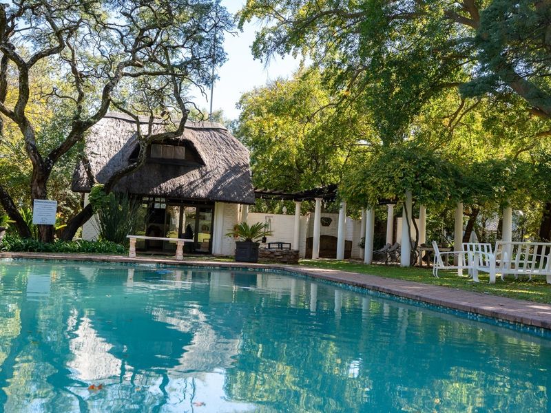 A serene outdoor pool area featuring a swimming pool, a charming thatched-roof structure, and a shaded pergola set within a lush, mature garden.