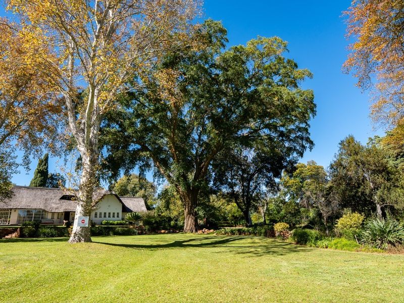 A spacious, well-maintained garden featuring mature trees and a view of a charming house with a traditional thatched roof.