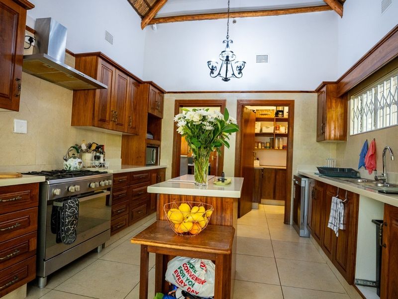 A well-maintained kitchen featuring warm wooden cabinetry, stone countertops, a gas stove with extractor, and exposed timber rafters, with an adjoining scullery area.