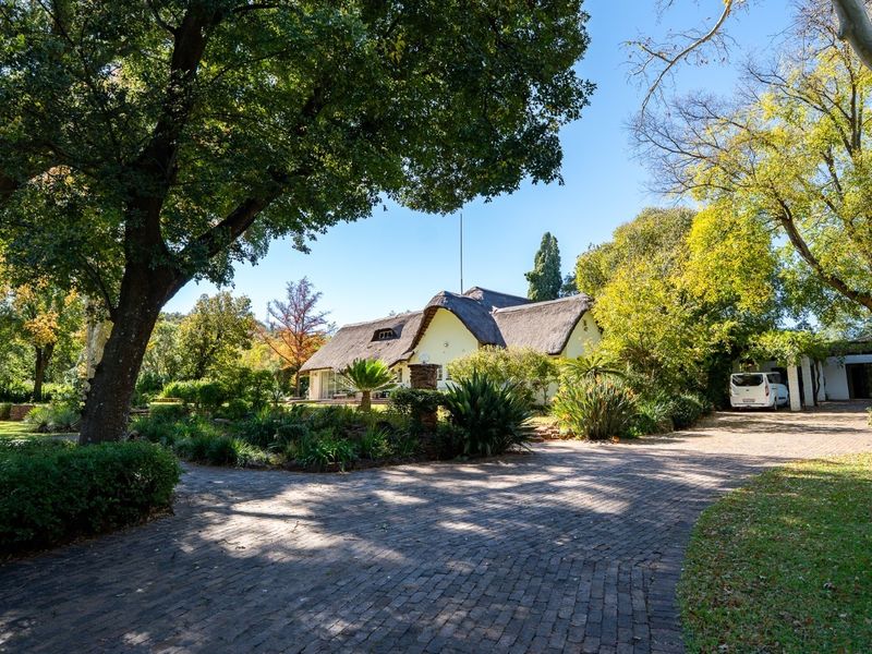 A charming thatched-roof house set within a lush, mature garden with a spacious brick-paved driveway leading to the entrance.