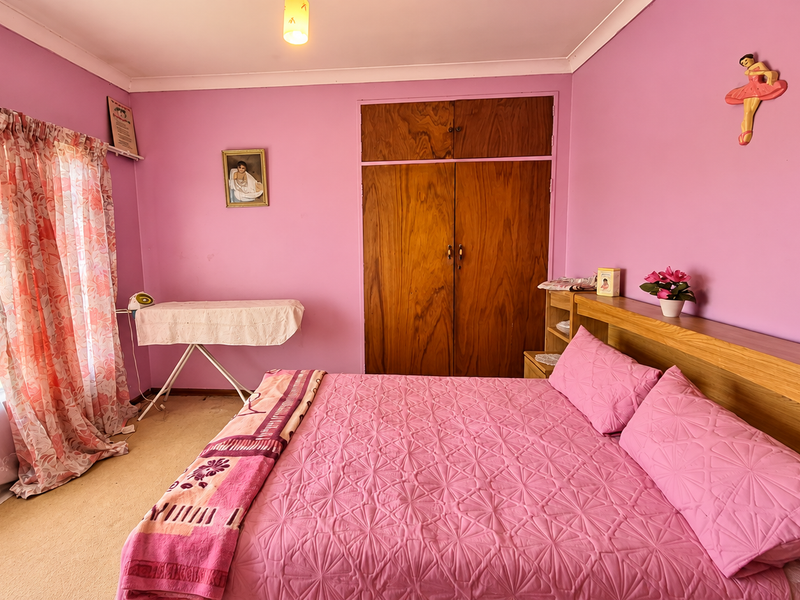 A bedroom featuring a built-in wooden wardrobe, integrated headboard, and large window with security bars, finished with carpeted flooring.