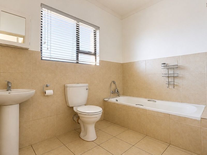 A functional bathroom featuring a pedestal basin, toilet, and bathtub with neutral wall and floor tiling and a window with venetian blinds.