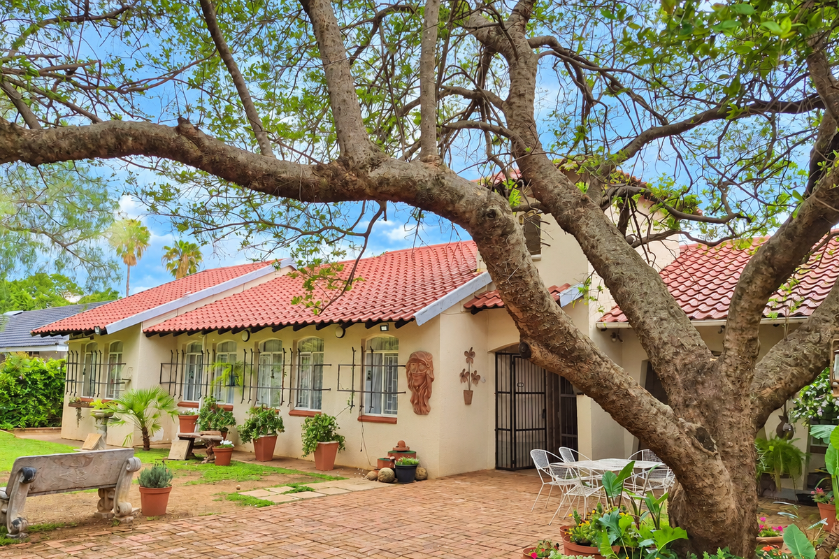 A charming rear exterior view featuring a terracotta tiled roof, brick paved patio, and mature trees providing shade over the garden area.