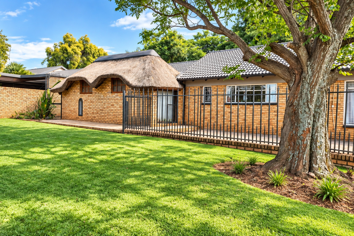 A well-maintained rear garden featuring a combination of thatched and tiled roof sections, brick exterior walls, and secure metal fencing.