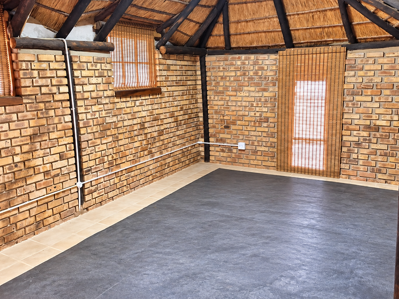 An empty living area featuring rustic exposed brick walls, a traditional thatched roof with timber beams, and a combination of tiled and dark flooring.